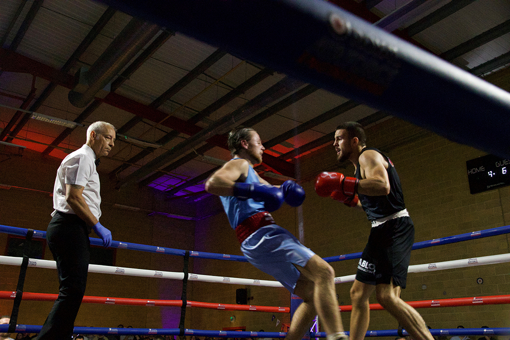 An action shot from a low angle looking up into a boxing ring, where a RAF boxer, in a light blue kit and blue gloves, trades punches with a boxer in a British Army, in black kit and red gloves. A referee in a white shirt watches the exchange closely.